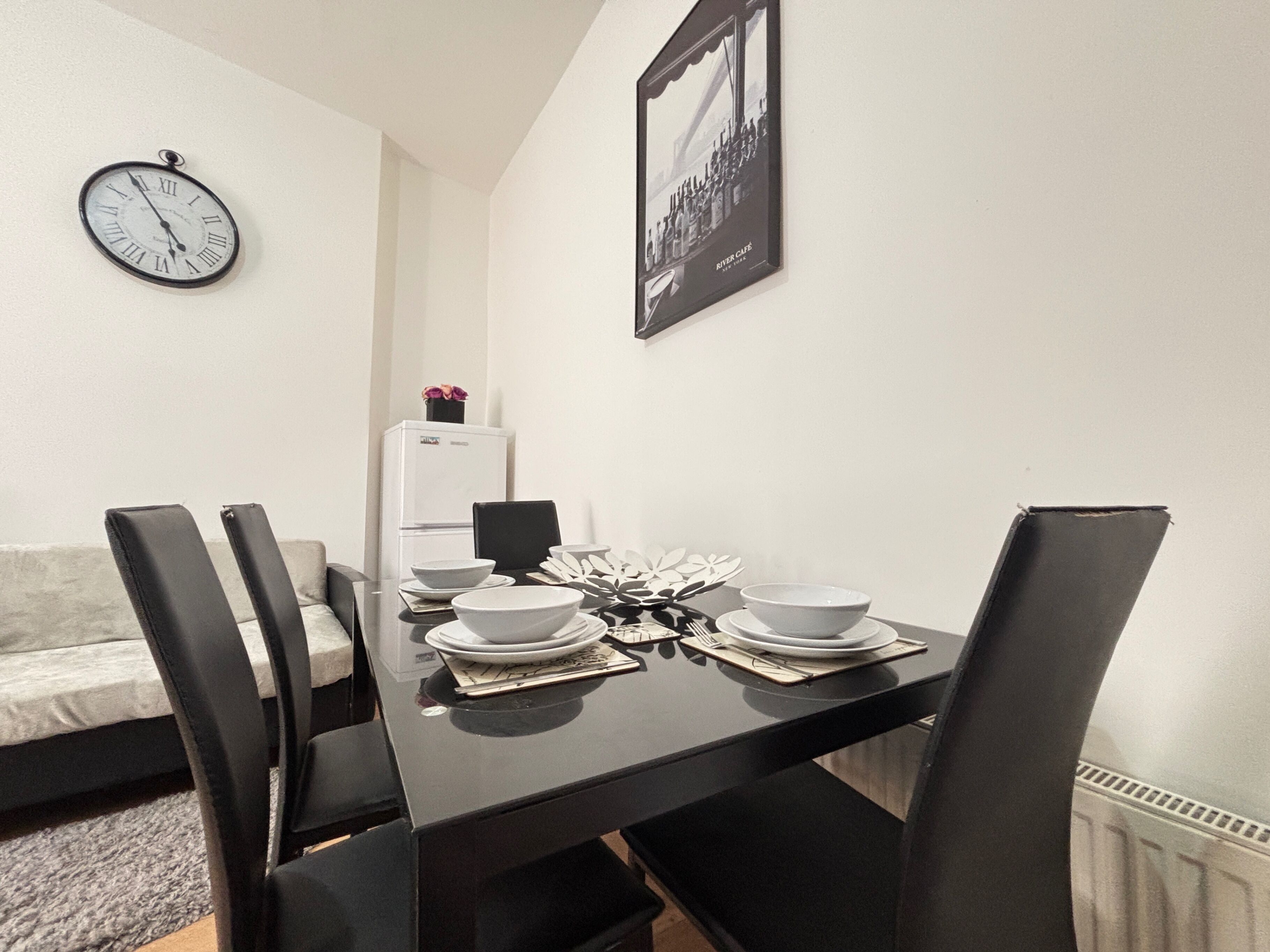Ein Essbereich mit einem schwarzen Tisch, gedeckt für vier Personen, weißen Tellern und einem schwarzen Stuhl. Im Hintergrund steht ein weißer Kühlschrank und an der Wand hängt eine Uhr.