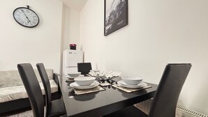 Een eetgedeelte met een zwarte tafel voor vier personen, witte borden en een zwarte stoel. Een witte koelkast staat op de achtergrond en een klok hangt aan de muur.