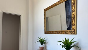 Een gang met een witte deur, een houten vloer, een witte console tafel met twee potplanten, en een grote, versierde spiegel aan de muur.