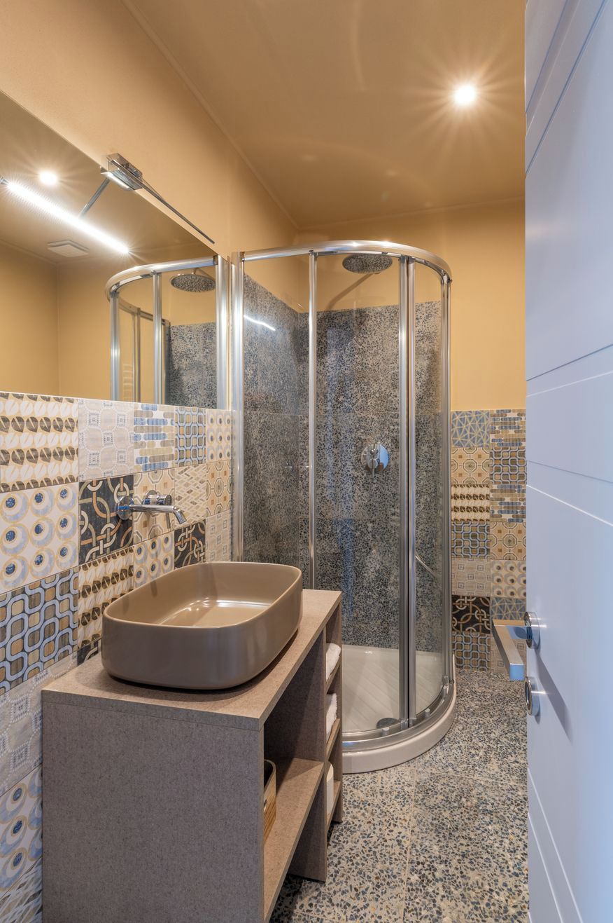 A modern bathroom with a curved glass shower enclosure, a built-in sink with a vessel basin, and patterned tiles on the walls and floor.