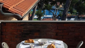 Una mesa con un croissant y una taza, frente a un muro de ladrillo con techo de azulejos y vistas a árboles y edificios.