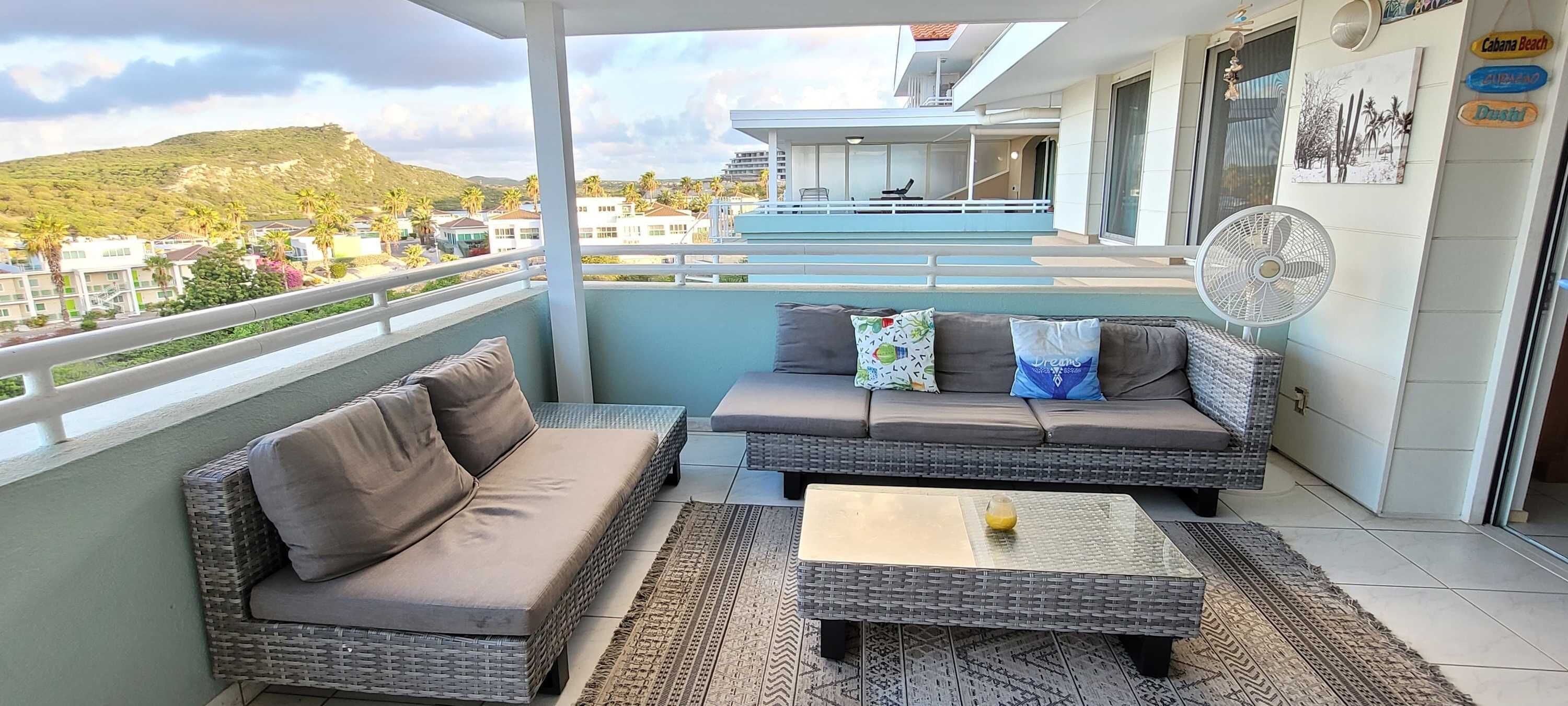 A balcony with wicker furniture, a fan, and a view of a building and palm trees.