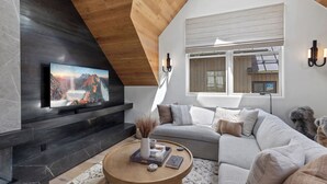 A modern living room with a large flat-screen TV mounted on a dark wall, a wooden ceiling, a grey sofa, and a round wooden coffee table with books and a vase.