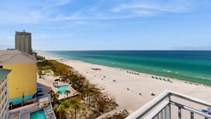 A beach view from a hotel balcony with a pool, beach chairs, and a tall building in the background.