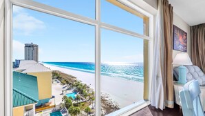 A hotel room with a large window overlooking a beach and a pool area.