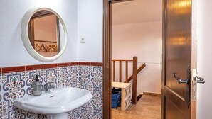 Ein Badezimmer mit einem weißen Waschbecken, einem Spiegel und gemusterten Fliesen. Eine Holztür führt zu einer Treppe.