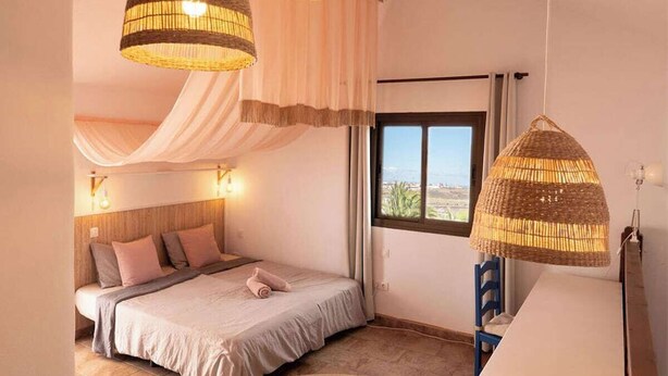 Ein Schlafzimmer mit einem Bett, zwei Hängeleuchten, einem Fenster mit Vorhängen und Blick ins Freie.