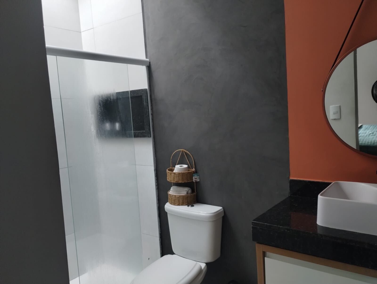 A bathroom with a white toilet, a glass shower enclosure, a dark gray wall, a round mirror, and a black countertop with a white sink.