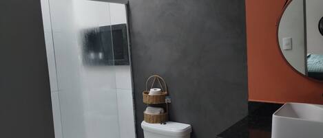 A bathroom with a white toilet, a glass shower enclosure, a dark gray wall, a round mirror, and a black countertop with a white sink.