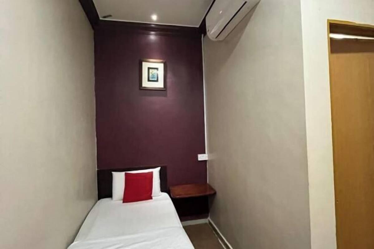 Ein kleines, schmales Hotelzimmer mit einem Einzelbett, einem roten Kopfkissen, einem gerahmten Bild an der Wand und einer hölzernen Bank.