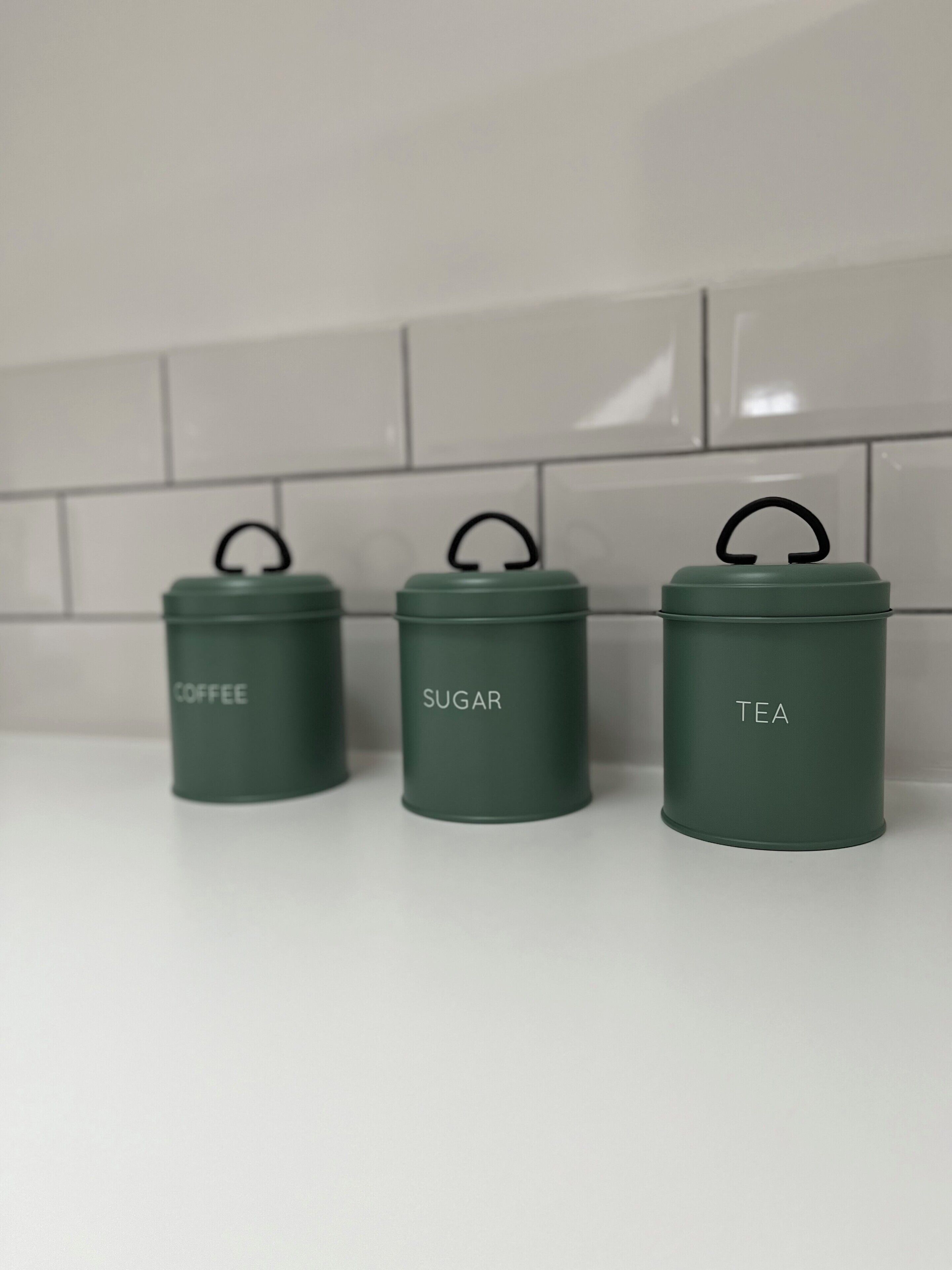 Trois récipients verts, étiquetés « café », « sucre » et « thé », sont posés sur un plan de travail blanc.