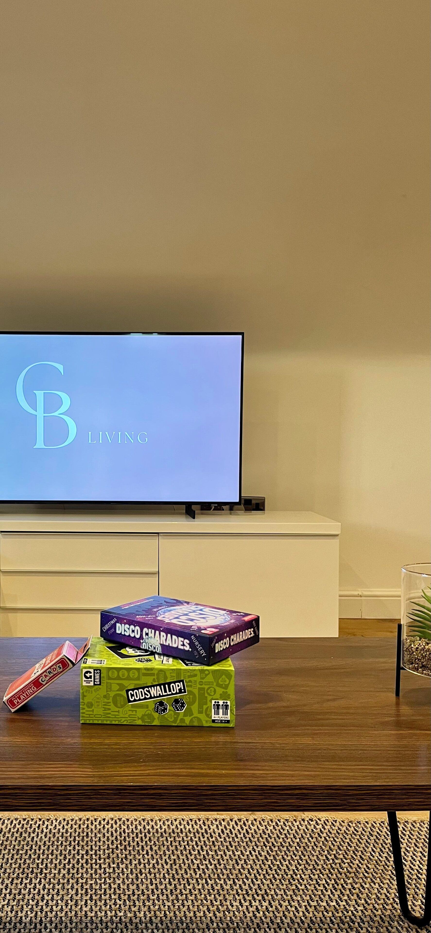 Una sala de estar con una televisión de pantalla plana sobre un mueble bajo, una mesa de centro con juegos de mesa y un florero de vidrio con plantas.