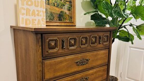 Un aparador de madera con varios cajones y tiradores decorativos, un cuadro enmarco con un paisaje, y un letrero con texto motivacional.