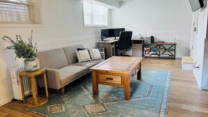 Una sala moderna con un sofá, una mesa de centro y una mesita auxiliar con una planta. Hay un escritorio con una computadora e impresora, y una estantería llena de libros.