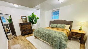 Un dormitorio con piso de madera, cama con ropa de cama verde y amarilla, cómoda, espejo, planta y ventana con persianas.