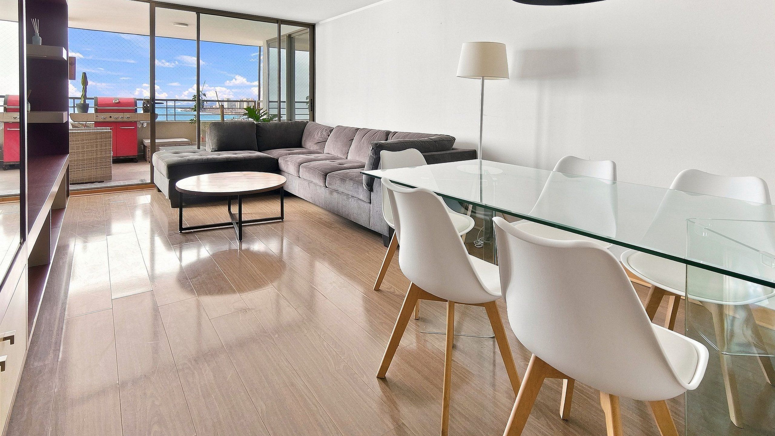 A modern living room with a glass dining table, white chairs, a grey sofa, and a large window offering a view of the sea.