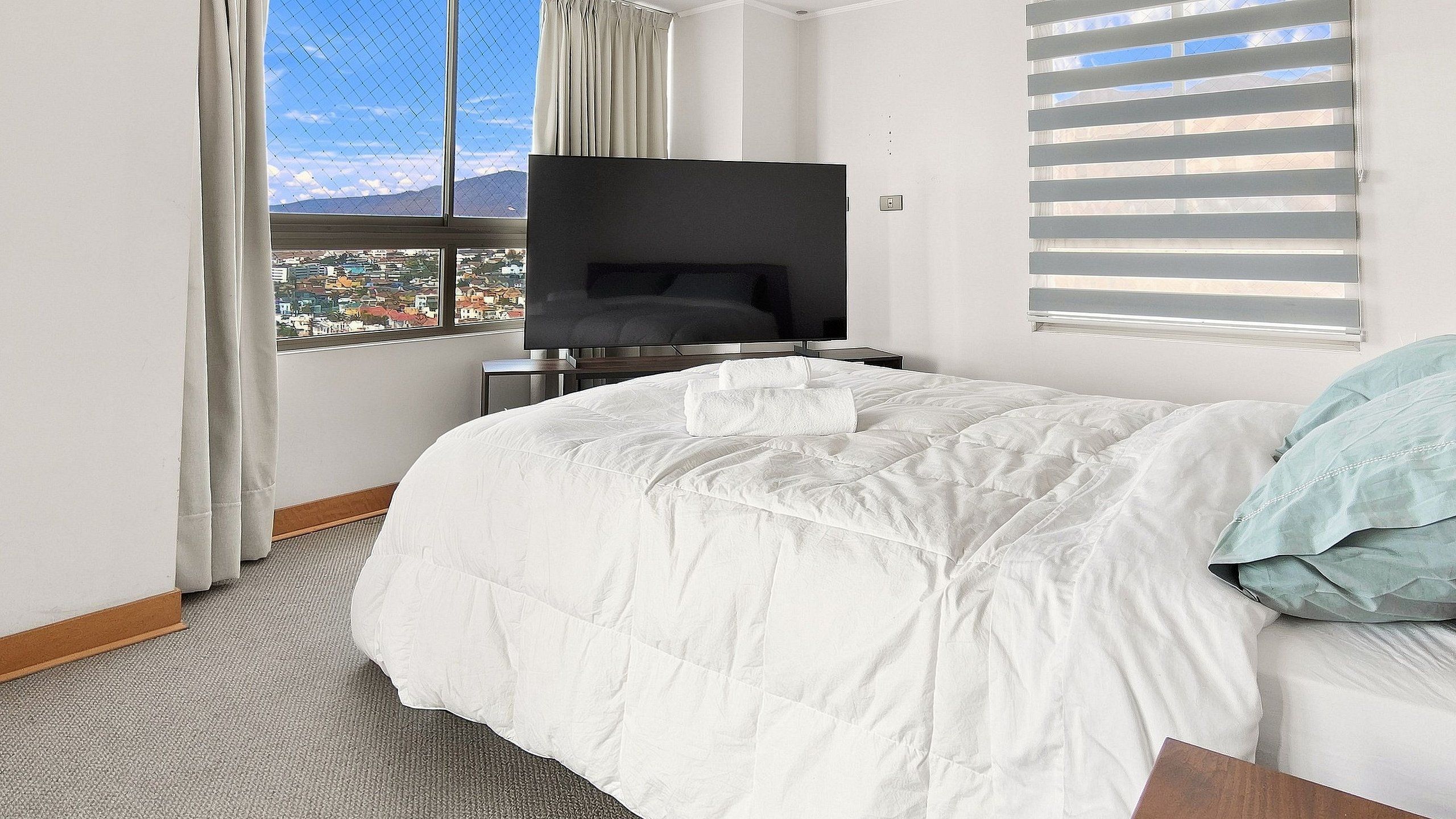 A hotel room with a large bed, a flat-screen TV, a city view, and a window with blinds.