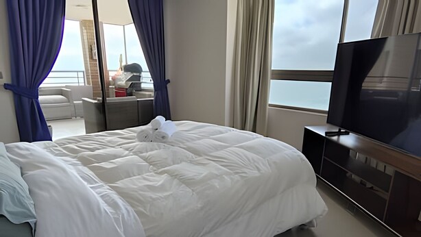 Ein Hotelzimmer mit einem großen Bett, einem Fernseher auf einem Holztisch und Meerblick durch das Fenster.