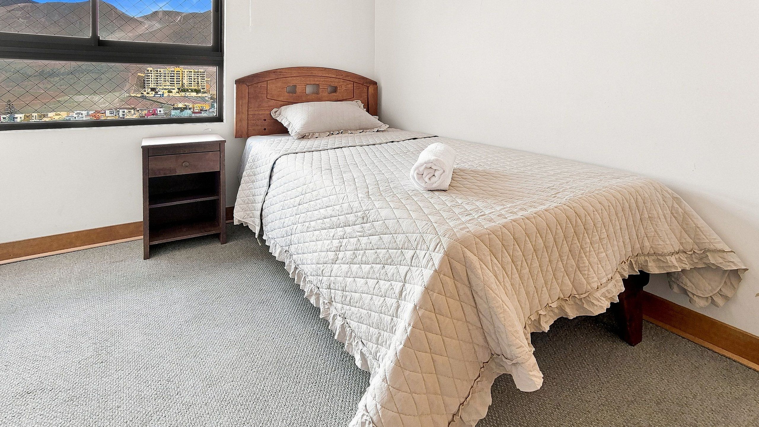 Un dormitorio con cama, mesita de noche, ventana con vistas a edificios y piso alfombrado.