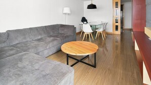 Ein modernes Wohnzimmer mit einem grauen Ecksofa, einem runden Couchtisch und einem Essbereich mit weißen Stühlen und einem Glastisch.