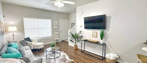 Una sala de estar con un ventilador de techo, un televisor de pantalla plana montado en la pared, una mesa de centro de madera y un sofá con cojines.