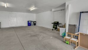 Una habitación amplia y vacía con piso de concreto, paredes blancas y una puerta. Hay dos botes de basura y una pequeña mesa con sillas.