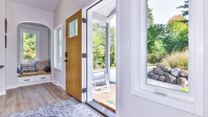 A modern interior with a wooden door leading to an outdoor deck, a bench with cushions, and a view of a garden with rocks and trees. - New Modern Construction on McNutt Lake, Minutes from downtown Minocqua! (Minocqua)