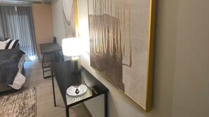 A modern hotel room with a large abstract painting, a desk with a lamp, a bed, and a view of the cityscape through the window. - Chill condo in San Fernando! (San Fernando)