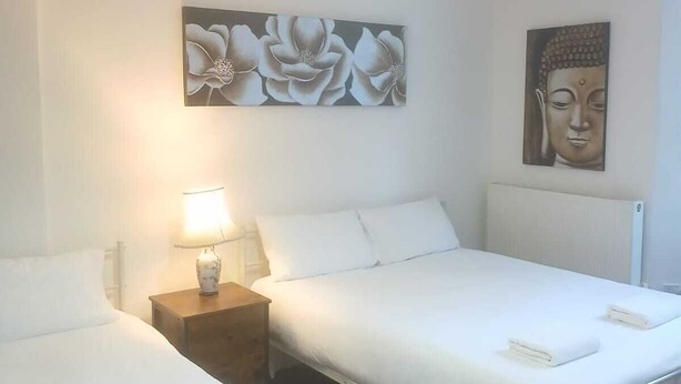 A bedroom with two beds, a nightstand with a lamp, and wall art. - Ground-Floor Family Studio Apartment with Double and Single Beds – Free Parking (Gosport)