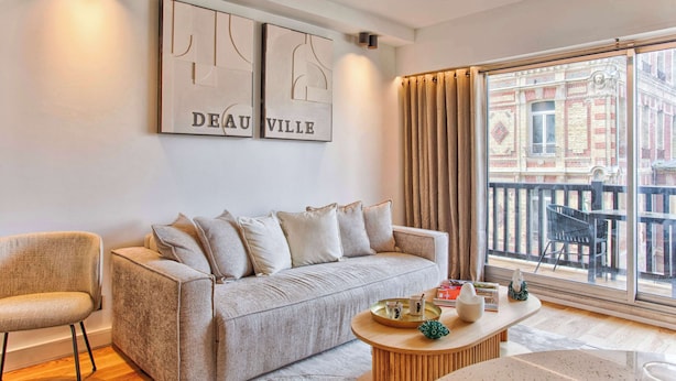 En moderne stue med en beige sofa, et rundt sofabord og et stort vindue med gardiner.