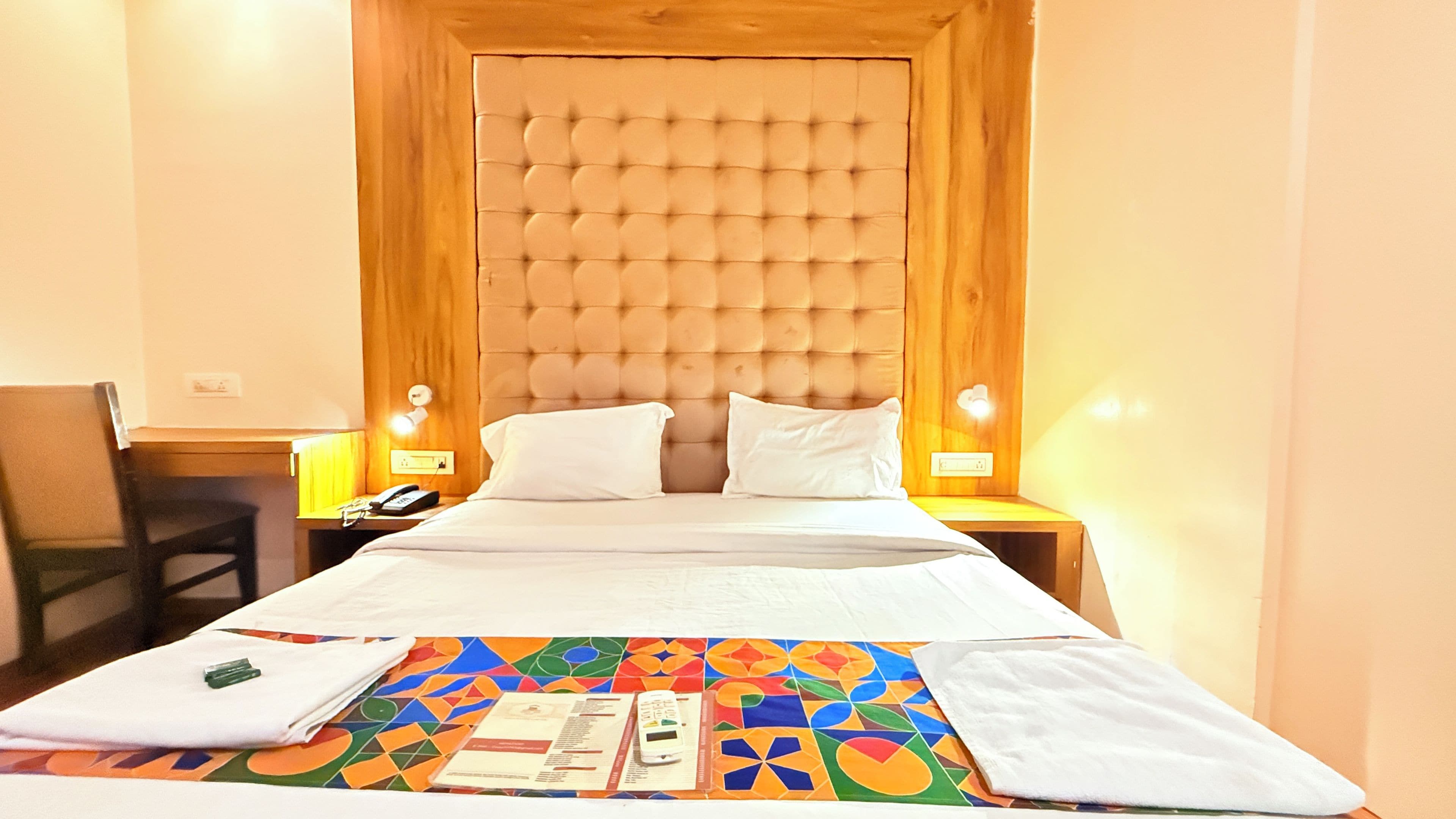 Une chambre d’hôtel avec un lit, une chaise, une table de chevet et une table agrémentée d’un chemin de table coloré.
