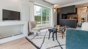 Ein modernes Wohnzimmer mit Fernseher, bequemen Sitzmöglichkeiten, Kamin und einem großen Fenster mit Blick ins Freie.
