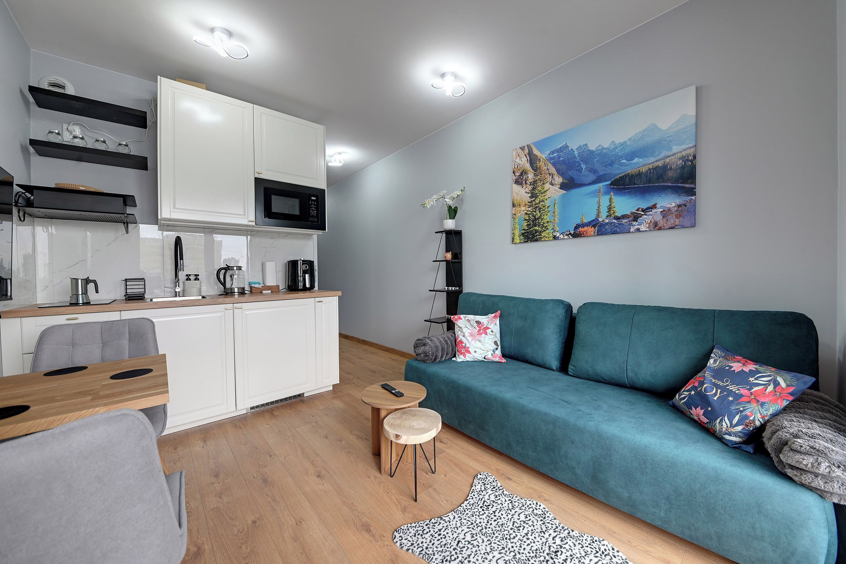 Una cocina moderna con armarios blancos, un microondas y una cafetera. Hay un sofá turquesa con almohadones decorativos y un cuadro con un paisaje montañoso en la pared.