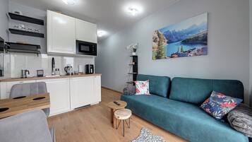 Una cocina moderna con armarios blancos, un microondas y una cafetera. Hay un sofá turquesa con almohadones decorativos y un cuadro con un paisaje montañoso en la pared.