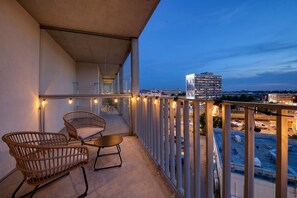A balcony with wicker chairs and a small table, overlooking a cityscape at dusk.