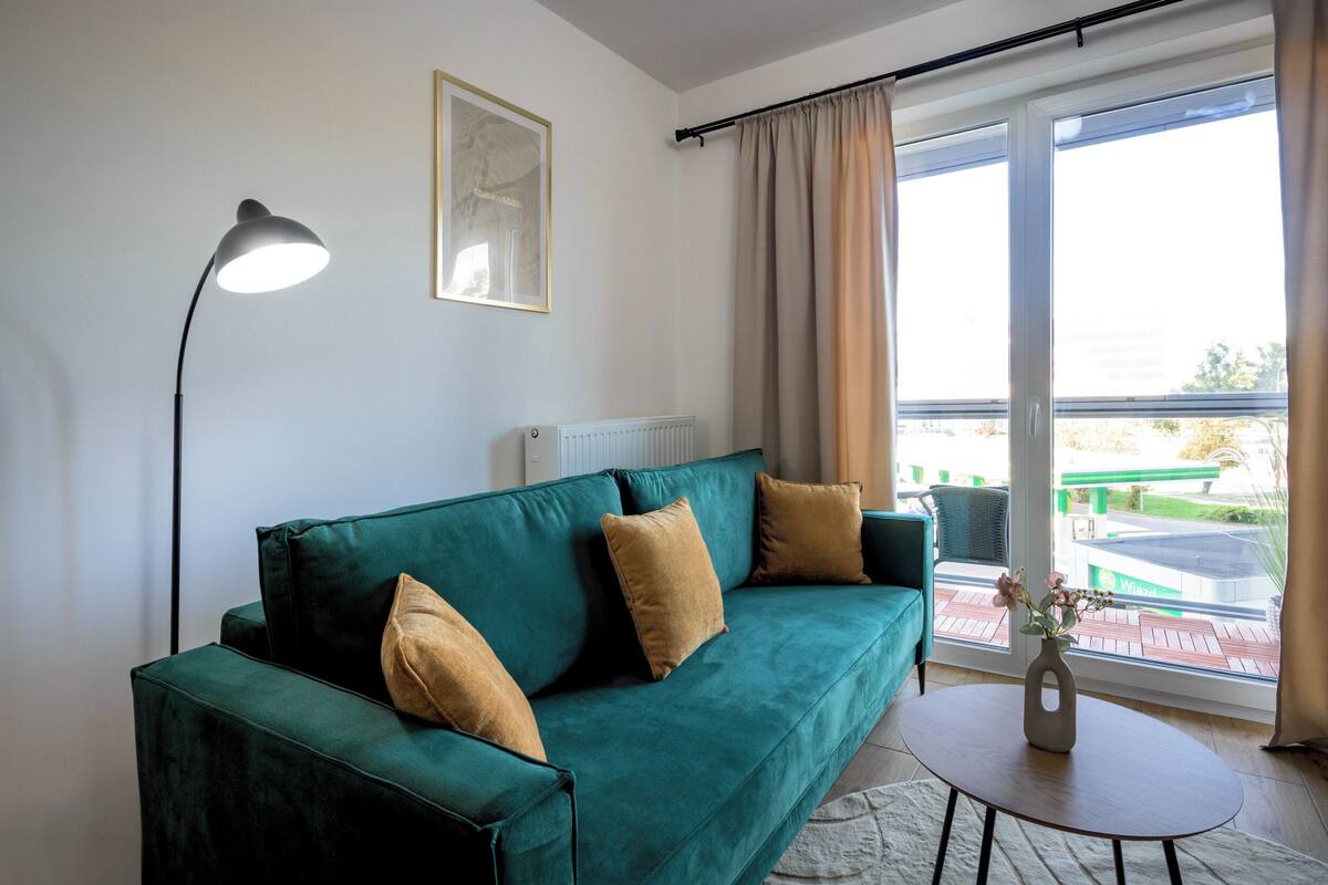 A green sofa with two cushions, a floor lamp, a small table with a vase, and a window with curtains.