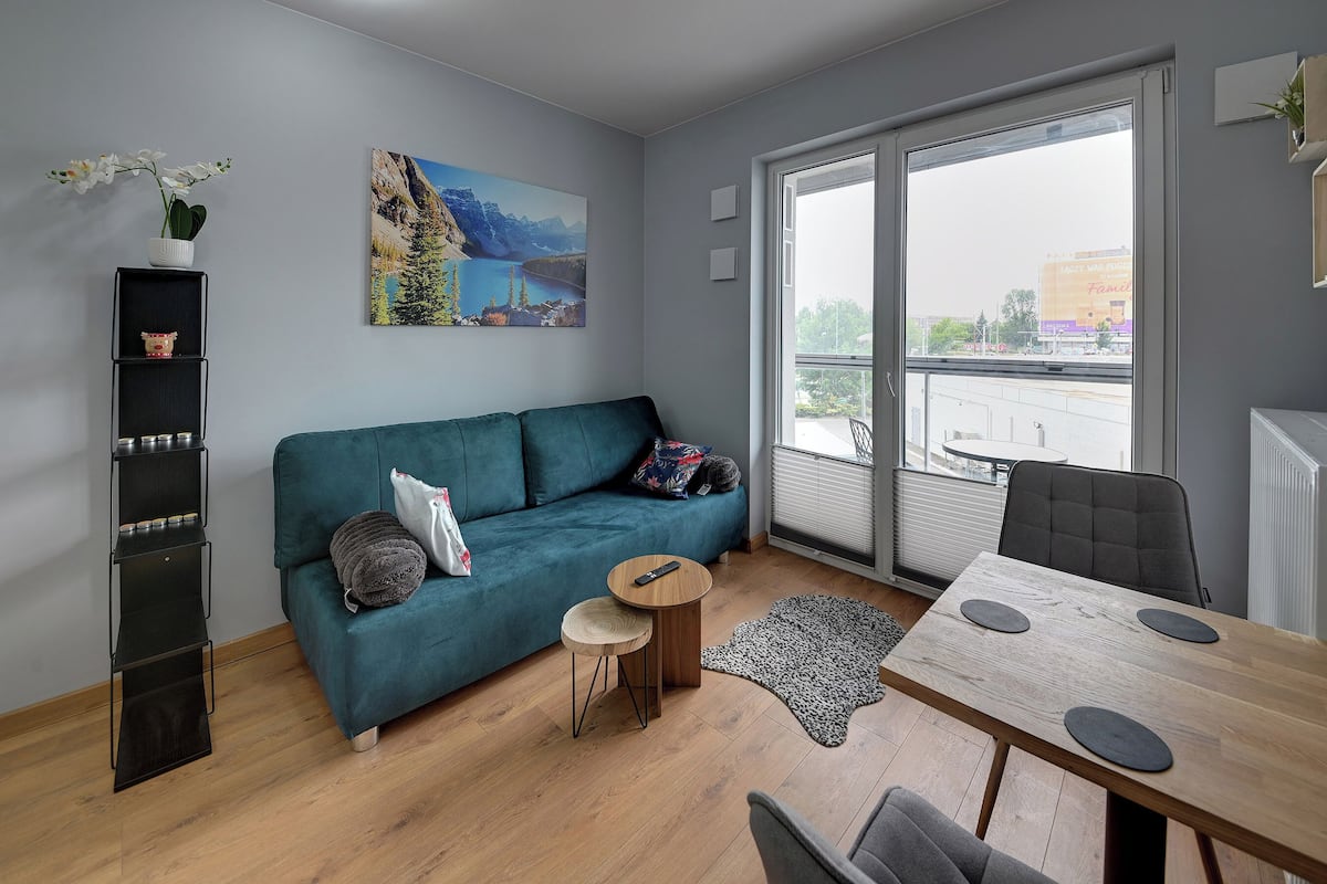 A modern living room with a teal sofa, a wooden dining table, a small side table, and a large window with a view of a building.