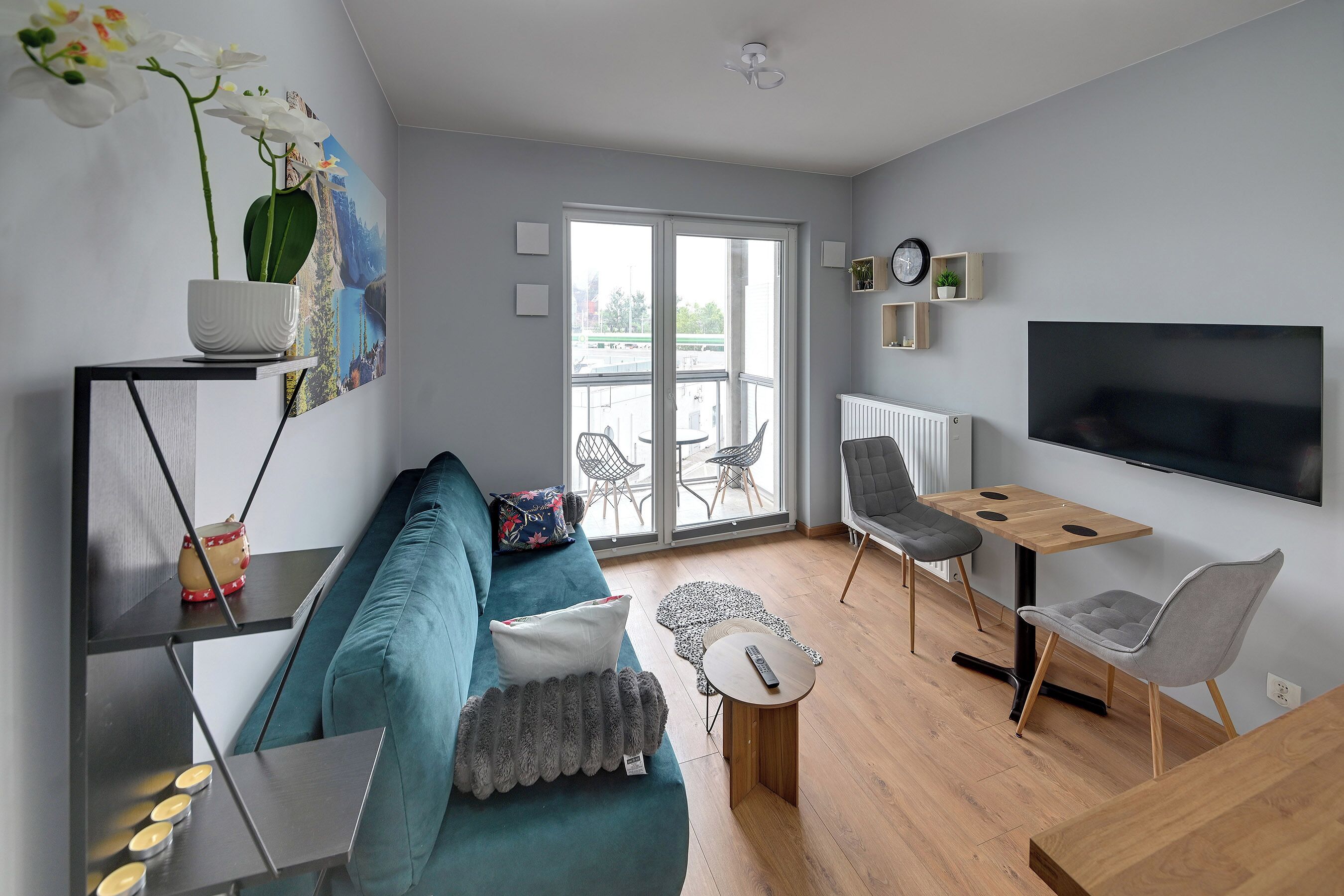 Una sala de estar moderna con un sofá color turquesa, una mesa de comedor de madera, un televisor y un balcón con mobiliario exterior.
