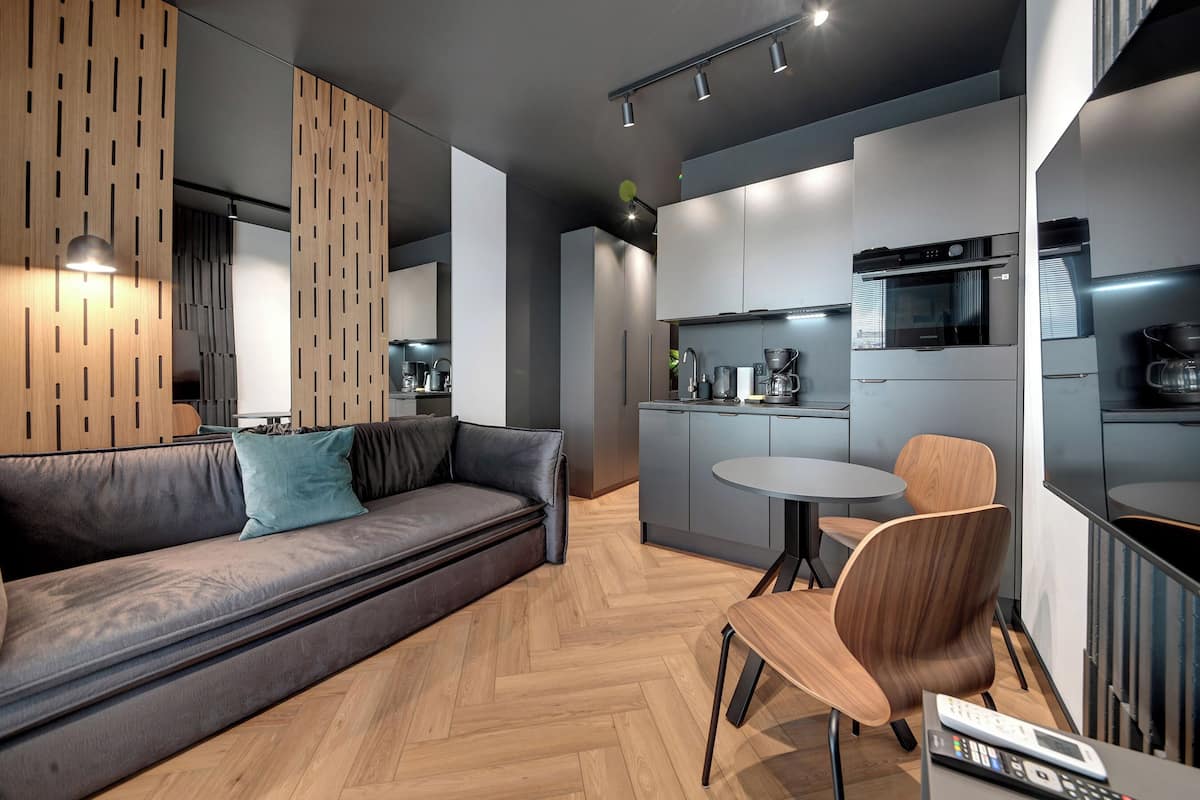 A modern kitchen with built-in appliances, a dining area with a round table and chairs, and a grey sofa with a teal pillow.