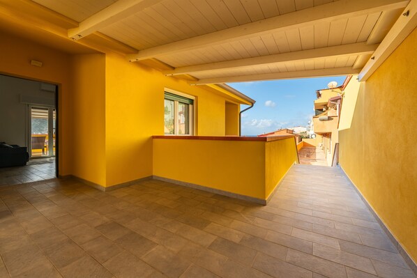A corridor with yellow walls and wooden flooring, leading to a balcony with a view of neighboring buildings.