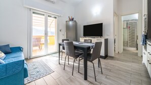Un espacio habitable compacto con comedor, sofá, televisor y puerta corrediza de vidrio que da a un balcón.