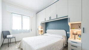 Un dormitorio con cama, una silla, una mesita de noche y una ventana.