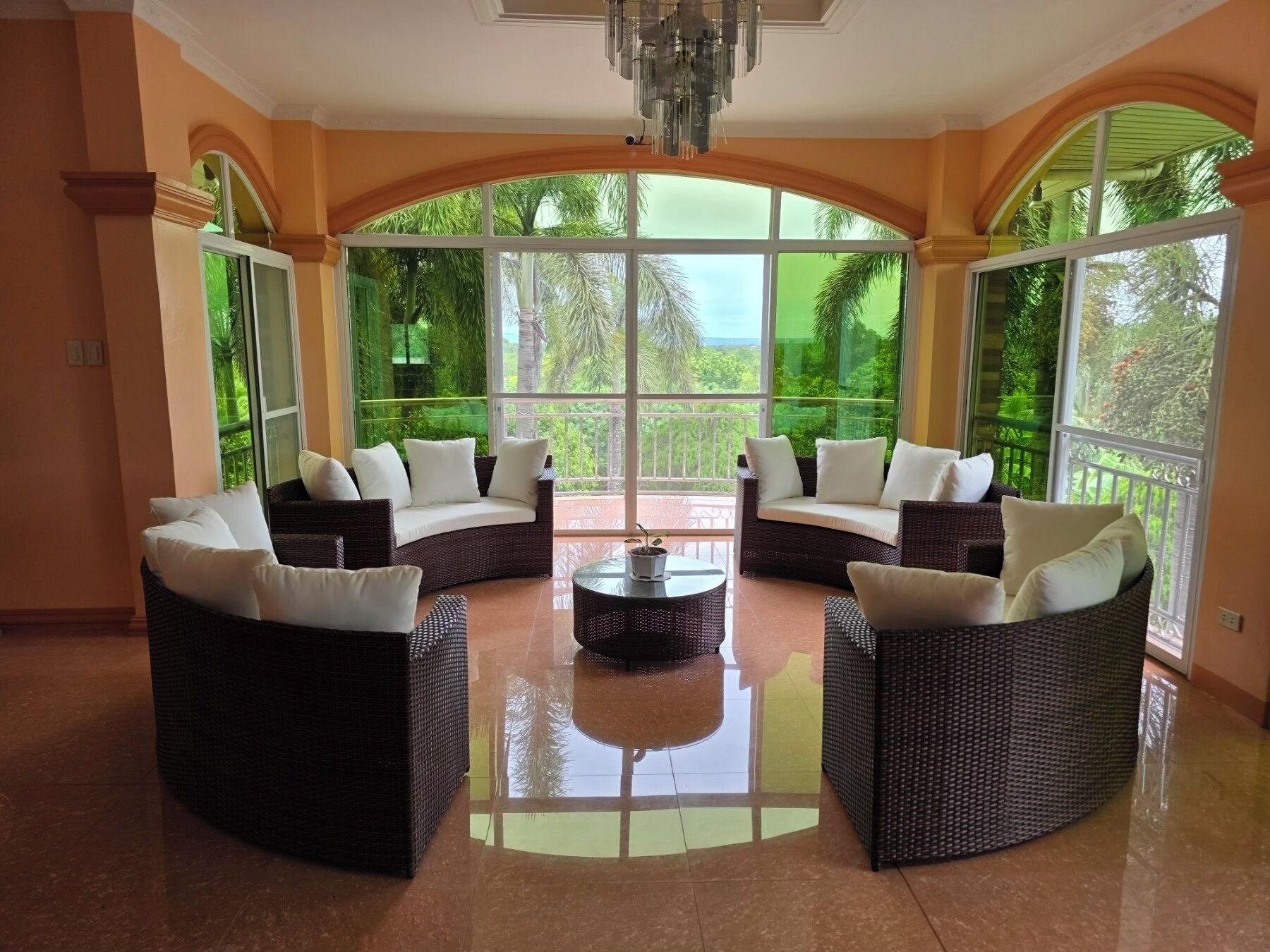 A spacious lounge with curved wicker furniture and a central round table, large windows offering a view of greenery.