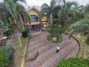 Un edificio de dos pisos con fachada amarilla y techo rojo, rodeado de palmeras y un patio con diseño.
