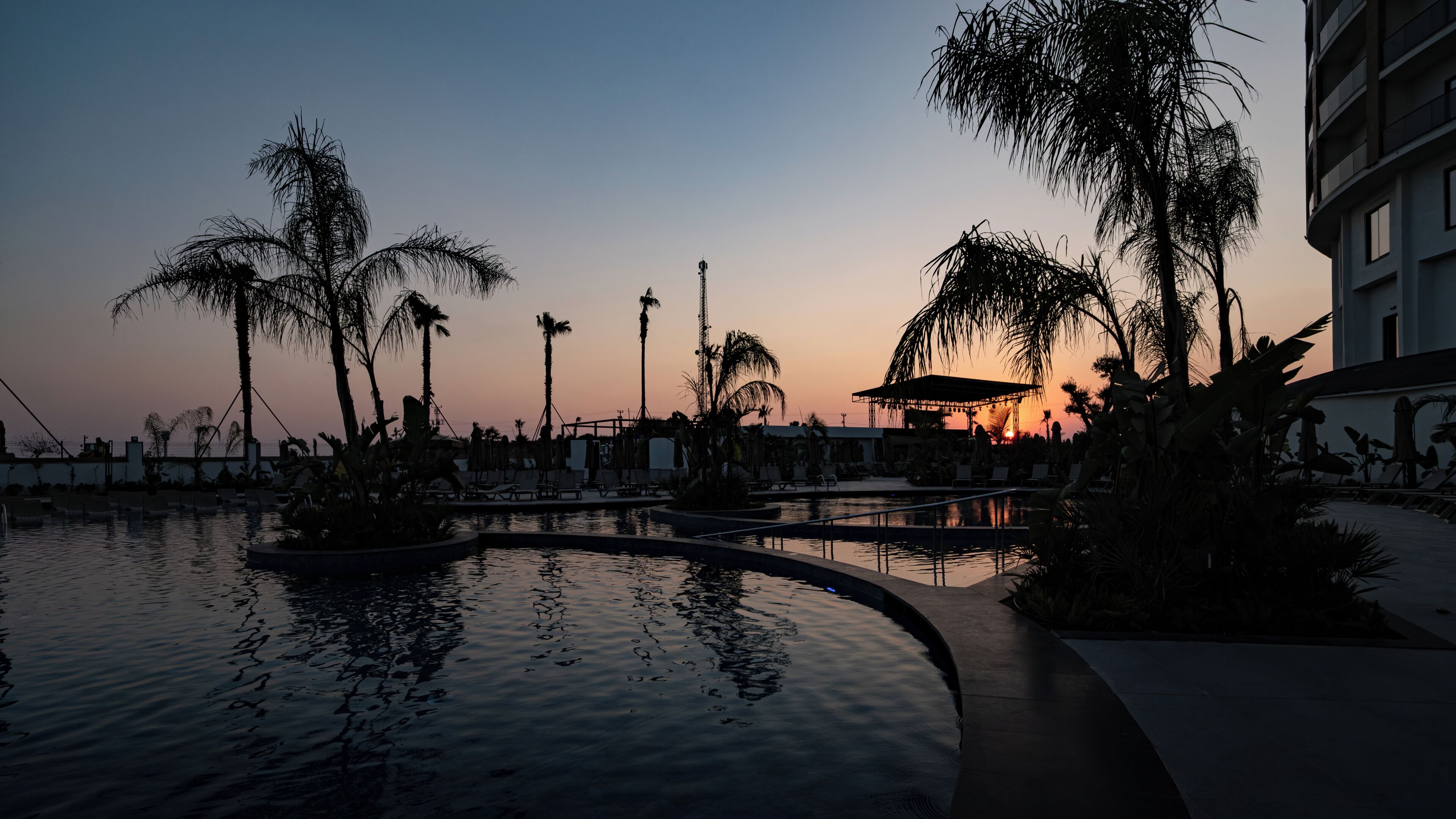 Zona de piscina en un resort, con palmeras y un atardecer de fondo.