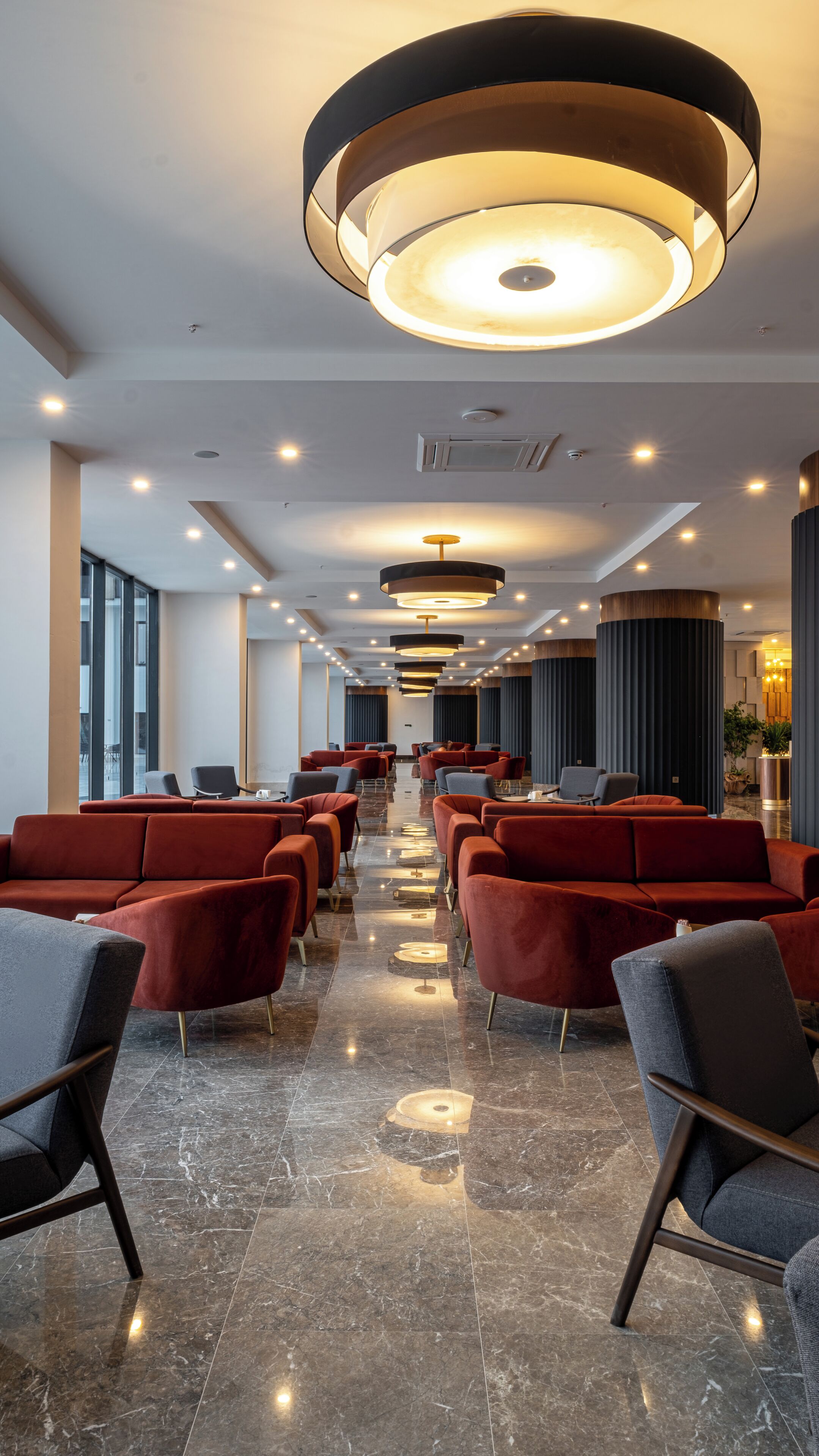 Un vestíbulo de hotel con hileras de sillones tapizados en rojo y gris, piso de mármol e iluminación colgante moderna.