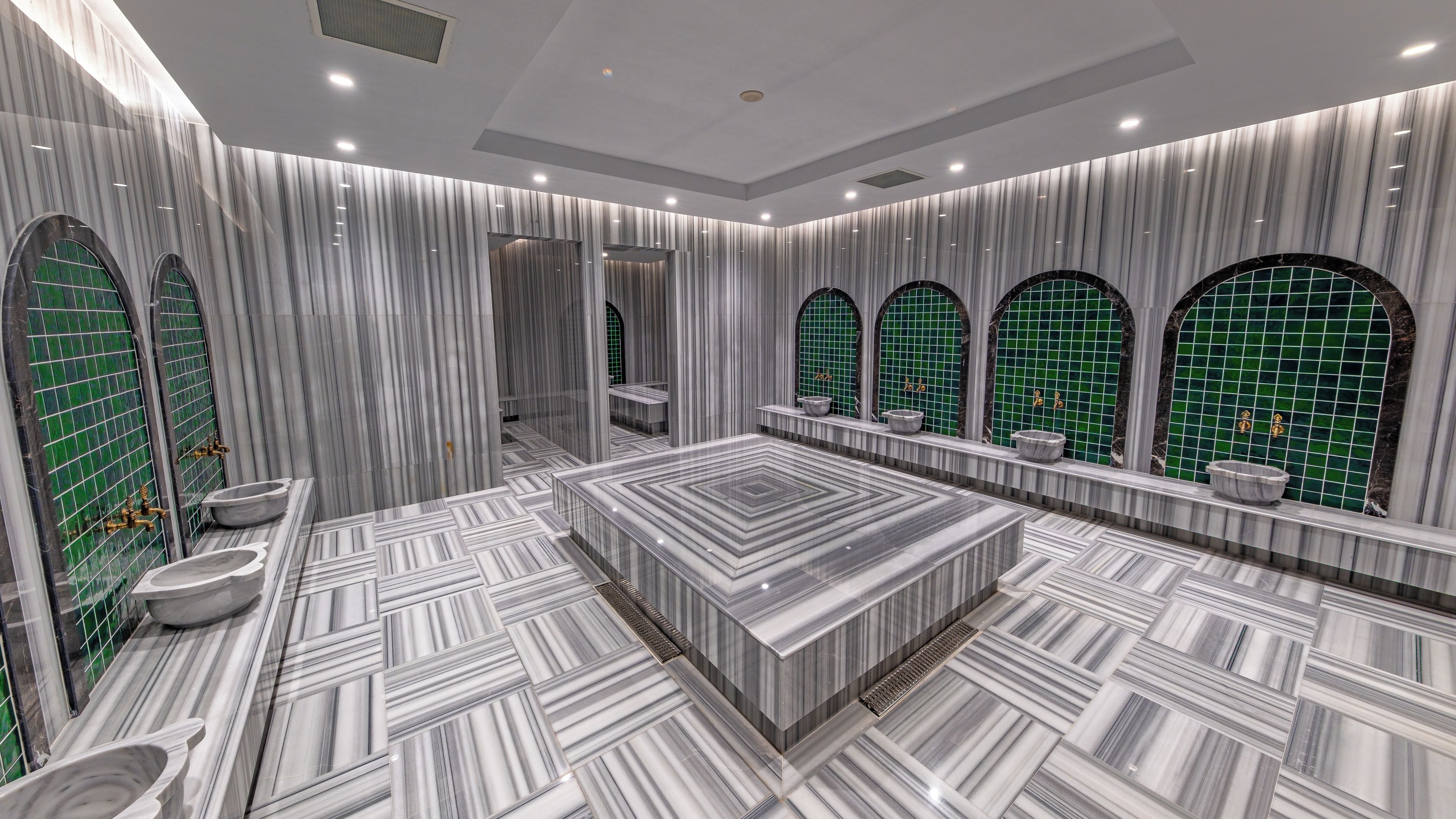 A modern bathroom with a marble floor, a large central sink, and green-tiled walls.
