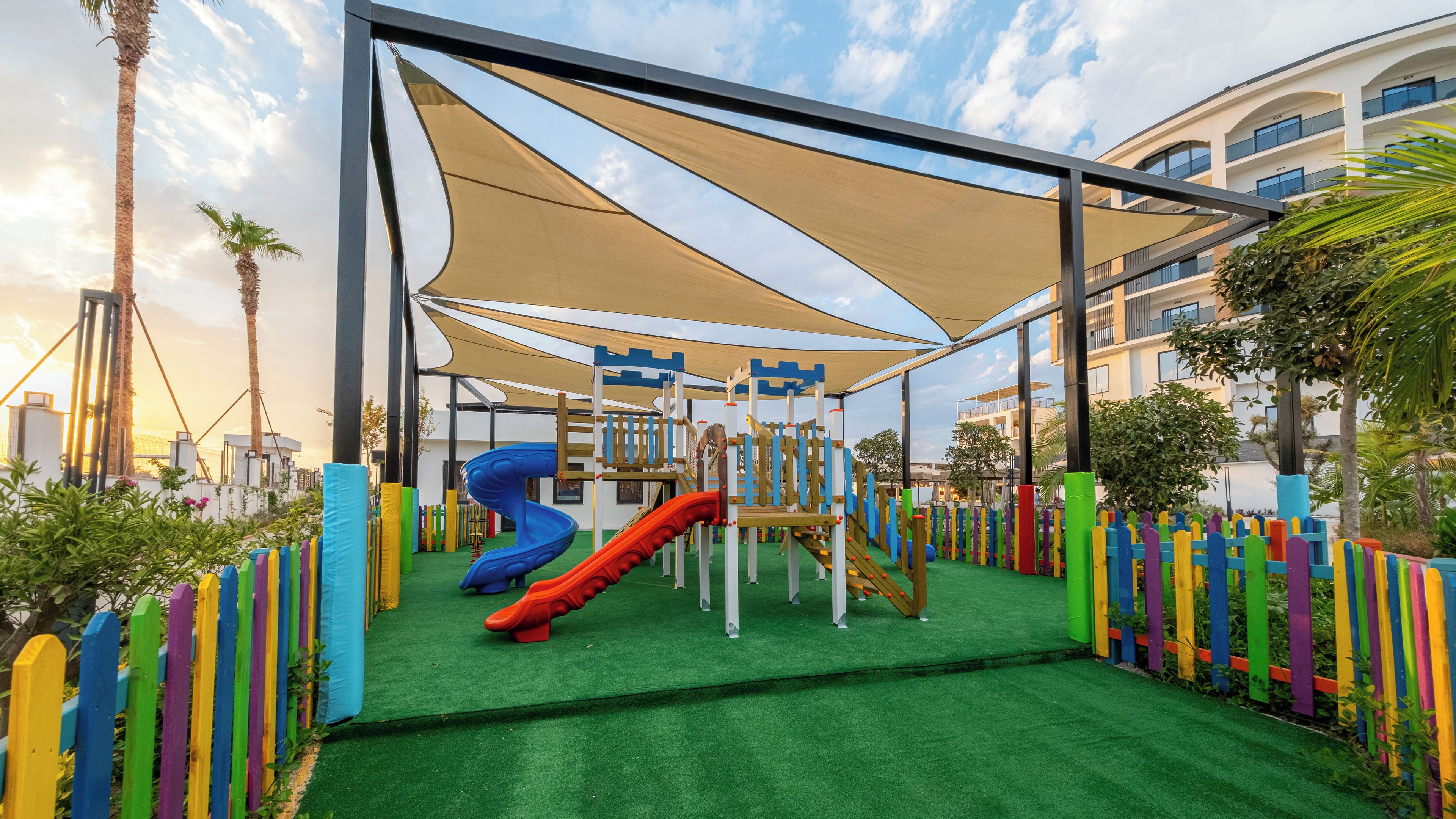 A playground with slides and climbing structures, surrounded by a fence and palm trees.