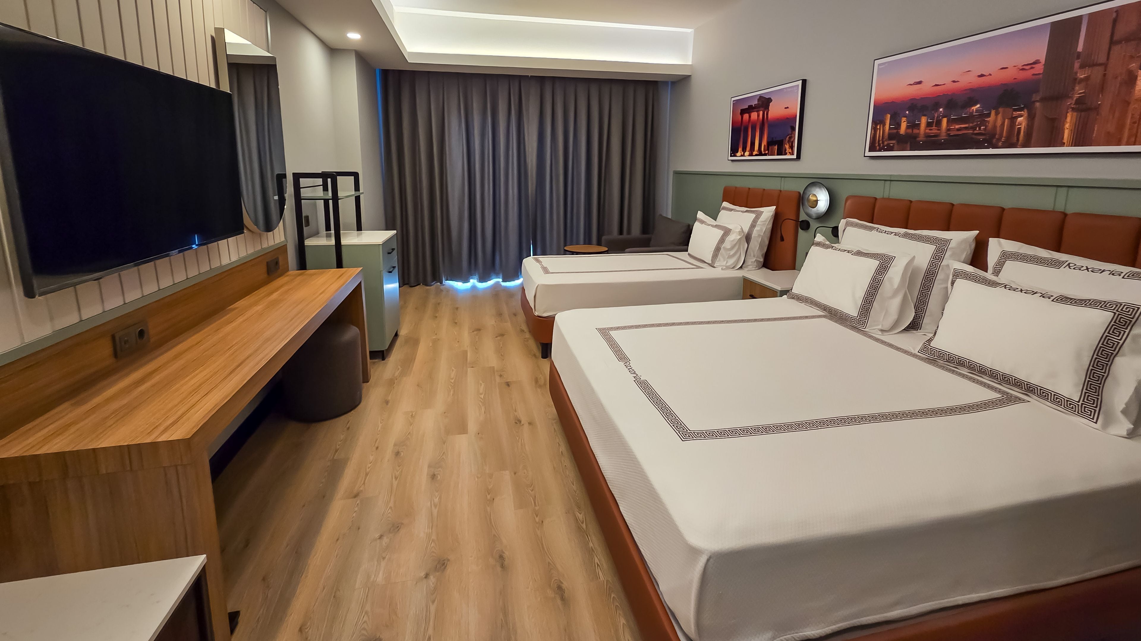 A hotel room with a TV, a desk, a bed with decorative pillows, a bedside table, and a large window with curtains.