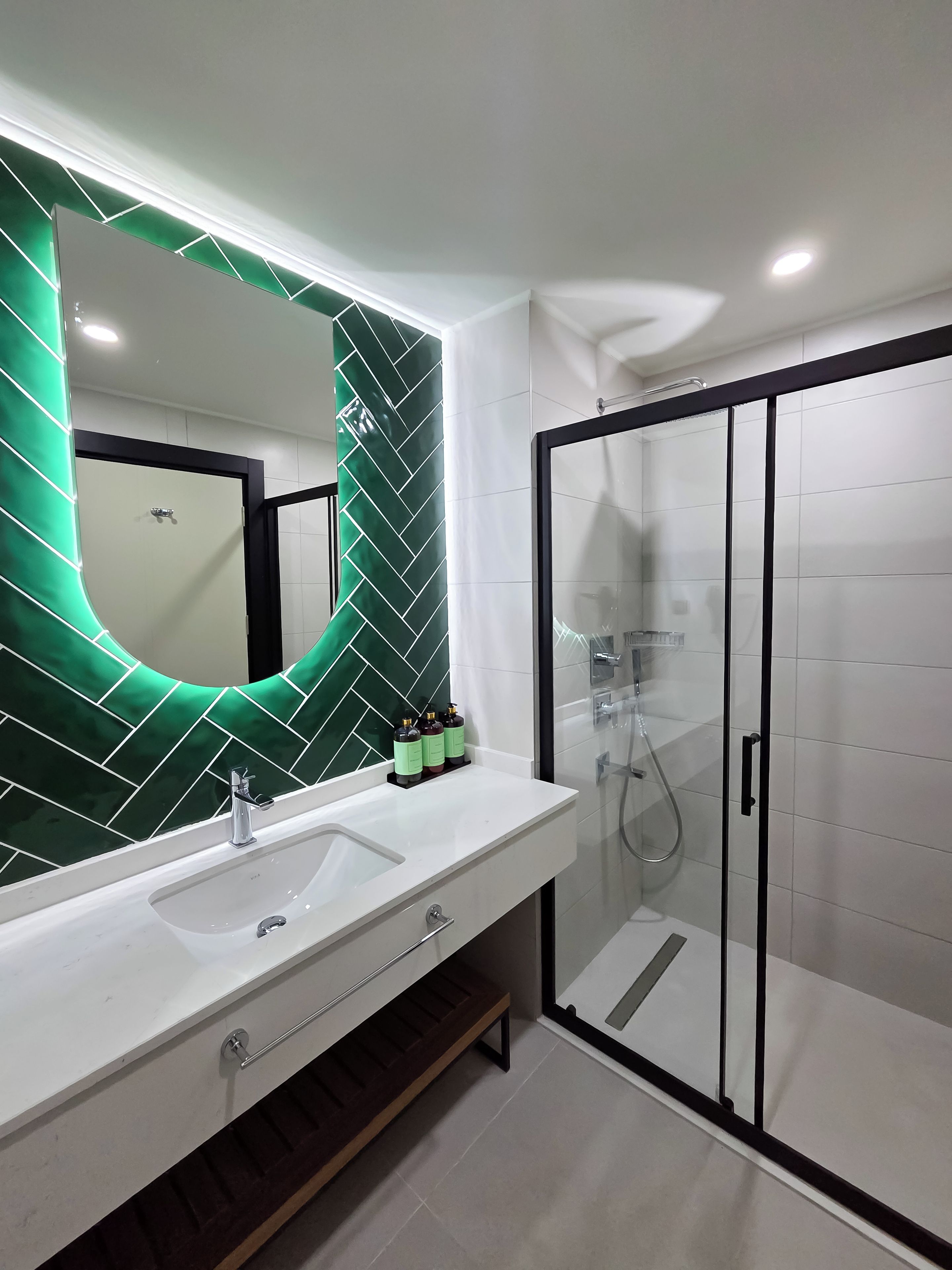 Un baño moderno con un lavamanos blanco, un espejo con un respuesto de estilo herringbone verde y una ducha con cerramiento de vidrio.